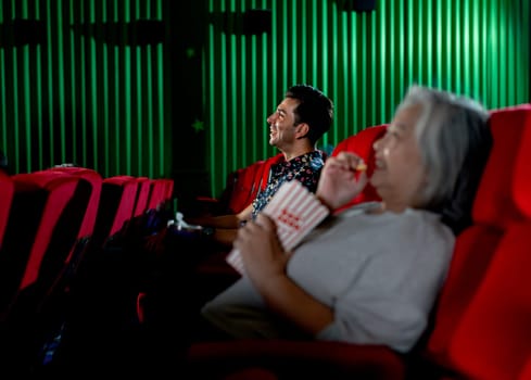 Caucasian man look happy with smiling during watch movie in cinema theater and senior woman also happy with eat popcorn as foreground.