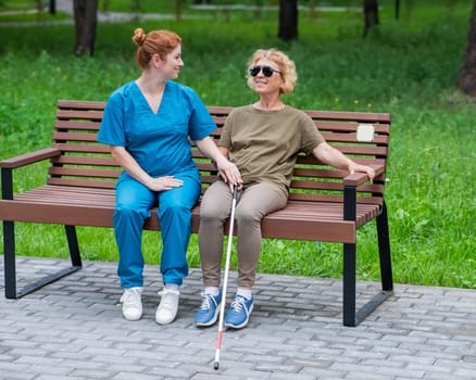 A nurse and an elderly blind woman are sitting on a bench in the park.