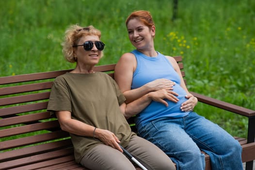 An elderly blind woman holds her pregnant daughter by the belly while sitting on a bench in the park.
