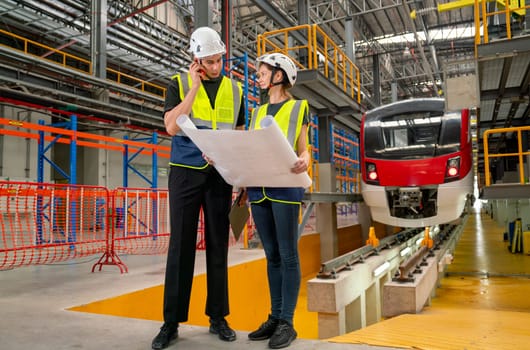 Wide shot of two professional engineer or technician workers man and woman discuss with drawing paper and stay in front of electric train in factory workplace.