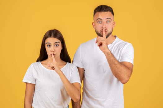 Surprised young couple in white t-shirts with fingers on lips making a "shush" gesture