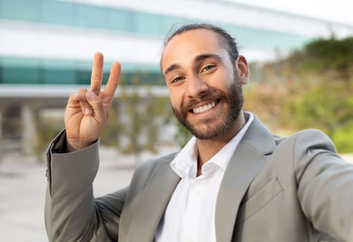 Friendly young businessman in suit making peace sign while taking selfie outdoors