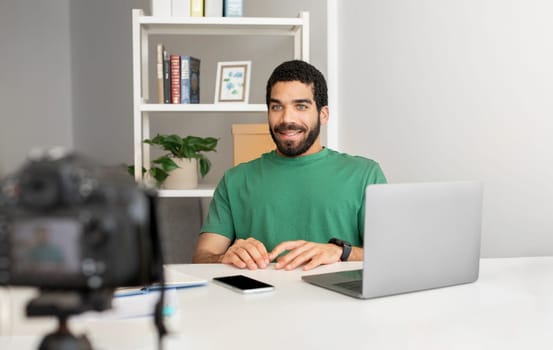 Smiling man in a green shirt sitting at a desk with a laptop, smartwatch