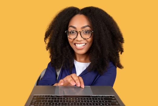 Cheerful black lady student typing at keyboard laptop and smiling on yellow background