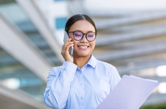 Japanese employee lady in eyewear speaking online on phone outside