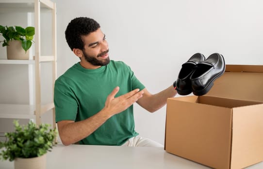 A cheerful young man in a green t-shirt enjoys unboxing a new pair of shiny black dress shoes