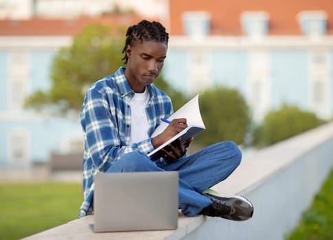 Black young man student browsing on laptop taking notes outdoors