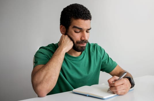 Serious man with a beard in a green t-shirt writing in a notebook, resting his chin