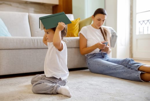 Preschool girl balancing book on head while her mother focused on smartphone