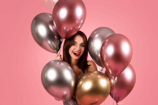 A delighted woman with dark hair and red lipstick smiles joyfully, surrounded by a cluster of metallic balloons