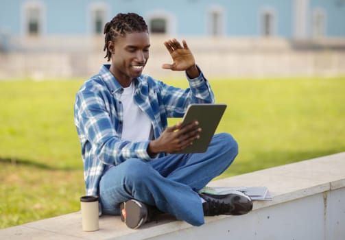 Happy black student guy waving hello to digital tablet outdoors
