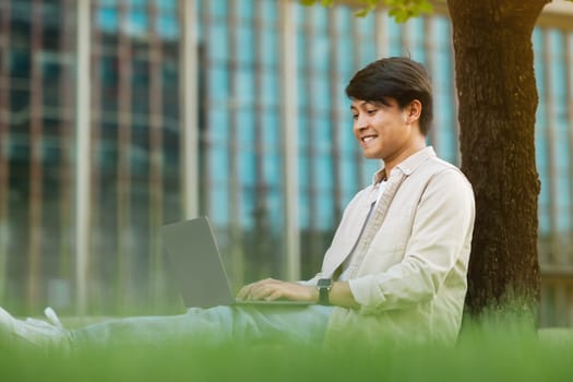 Asian guy freelancer sitting under tree, working on laptop