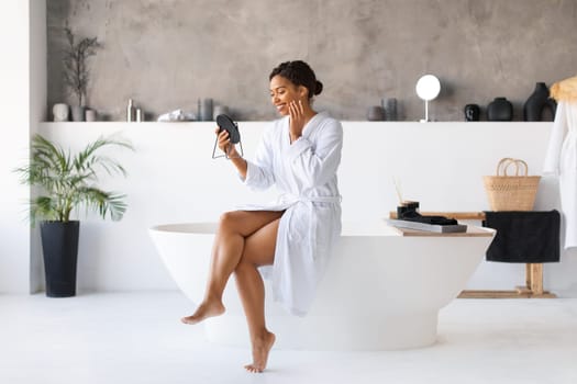 Attractive young black woman sitting on bathtub and looking into handheld mirror