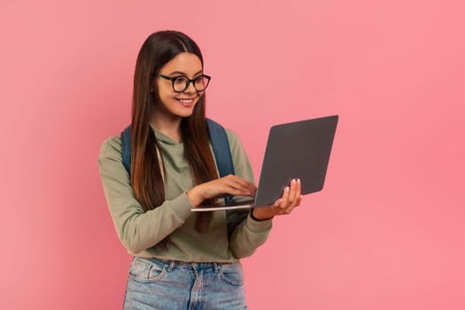 Smiling teenage girl wearing eyeglasses and backpack looking at laptop screen