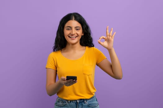 Cheerful attractive young hindu lady with smartphone showing okay