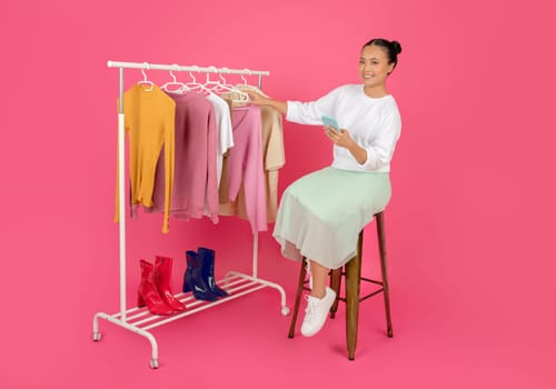 Asian woman with smartphone in hand comfortably seated near clothing rack