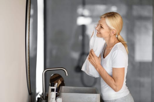 Happy mature woman in casual clothes drying face with soft white towel