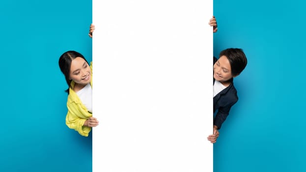Asian man and woman playfully peeking around a large white vertical banner