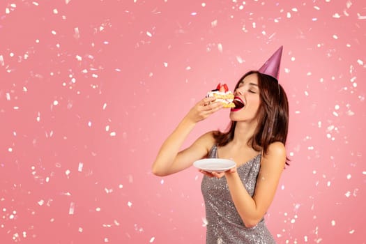 Cheerful pretty millennial caucasian brunette woman in party cap, dress eat cake