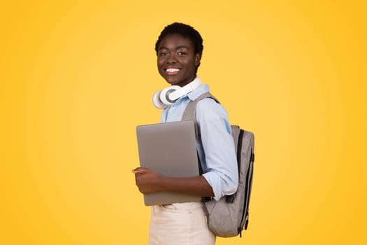 Smiling teen african american lady student in wireless headphones, hold laptop, ready to lesson