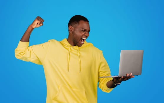An exuberant Black man in a yellow hoodie raises his fist in victory as he looks at a laptop