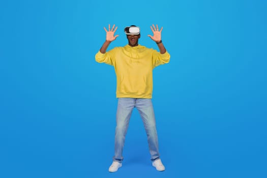 A Black man in a yellow hoodie stands amazed with hands raised, wearing virtual reality glasses
