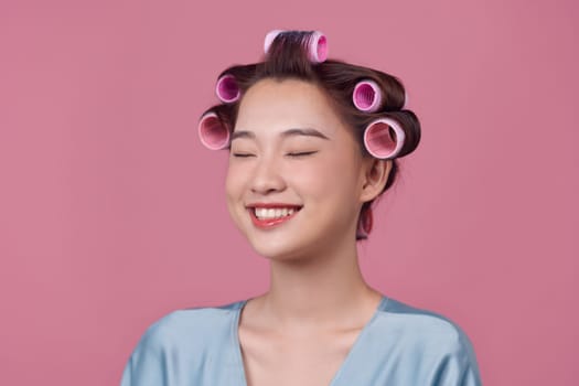 Beautiful young woman in silk dress with hair curlers on pink background. Space for text