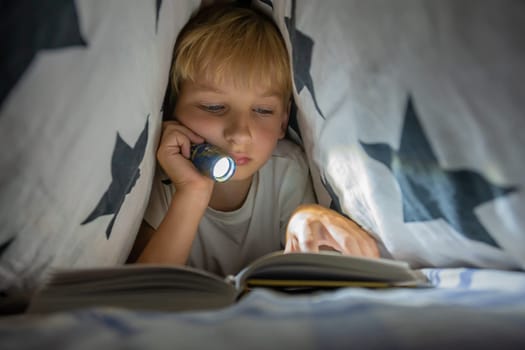 A little boy reads a book with a flashlight under the covers at night.