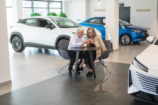 A mature couple is sitting in a car dealership and looking at a smartphone.