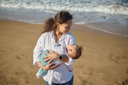 Authentic portrait of a happy family of a beautiful young mother in white casual shirt and blue jeans, carrying her beloved baby boy while walking by ocean on the beach in summer. Mom and son together