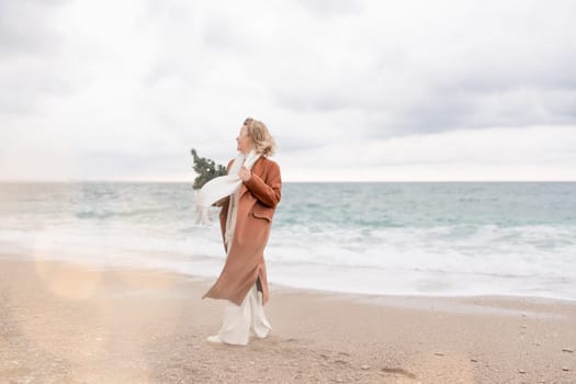 Blond woman Christmas sea. Christmas portrait of a happy woman walking along the beach and holding a Christmas tree in her hands. She is wearing a brown coat and a white suit