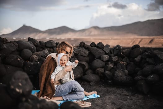 Mother enjoying winter vacations playing with his infant baby boy son on black sandy volcanic beach of Janubio on Lanzarote island, Spain on windy overcast day. Family travel vacations concept.