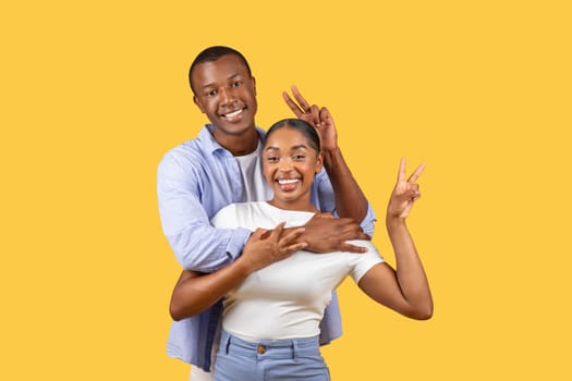 Playful couple making peace signs, joyful embrace, yellow background