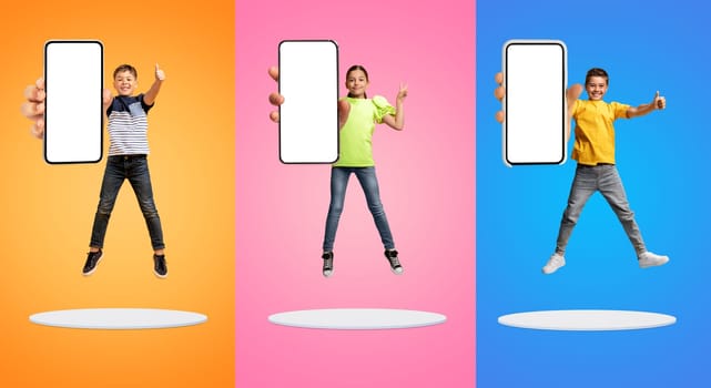 Happy kids showing thumbs up while jumping with blank-screen smartphones in hands