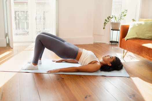 Young fit lady practicing yoga doing bridge pose on fitness mat in modern living room at home. Woman in fitwear focusing on daily workout training and muscles flexibility in the morning