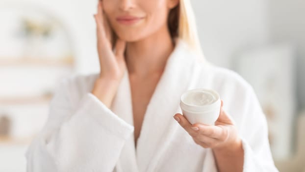 Cropped view of blonde lady holding jar of moisturizer indoor