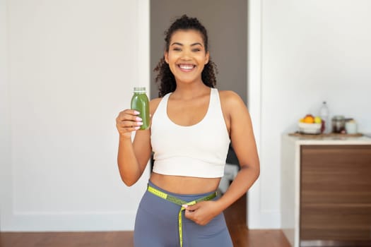 lady holding green smoothie and measuring waist with tape indoor