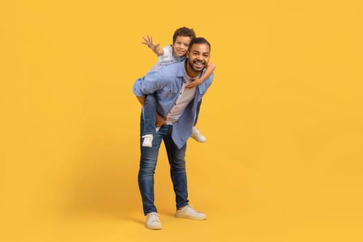 Loving black dad giving his son a piggyback ride over yellow background