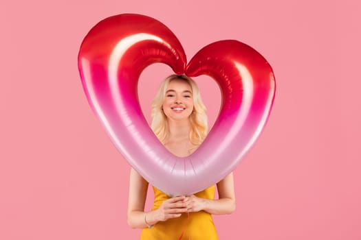 Smiling blonde woman with heart-shaped balloon on pink background