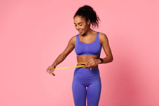 Excited black fitness lady measuring slim waist with tape, studio