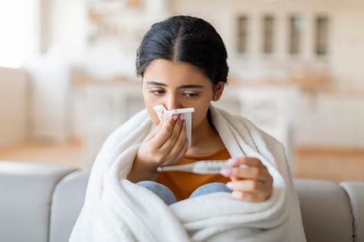 Sick Indian Woman Blowing Runny Nose And Looking At Thermometer At Home