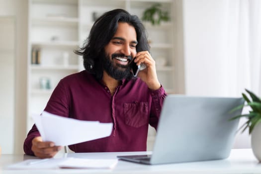 Indian man discussing work on phone while reviewing documents and using laptop