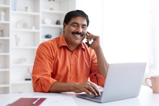 Middle aged eastern businessman talking on cellphone with client or business partner, sitting at desk at home office, typing on computer keyboard and smiling, giving consultation on phone