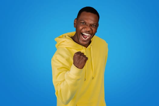 An exuberant African man in a yellow hoodie punches the air with a clenched fist
