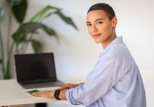 A professional latin smiling young woman with short hair confidently poses by a desk with a laptop, embodying modern workplace elegance and efficiency, enjoy work, study