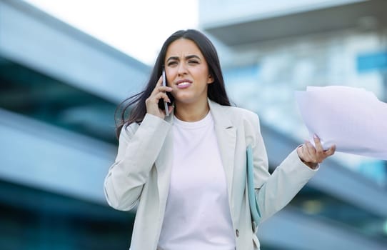 Angry business lady speaking on smartphone holding important documents outdoors