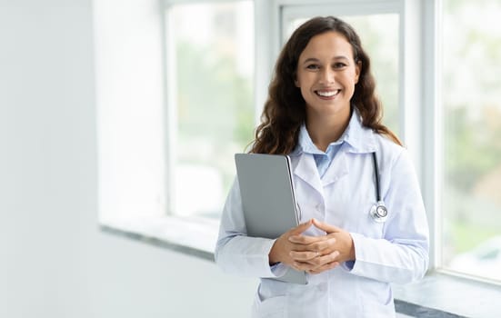 Woman doctor standing by window, holding laptop