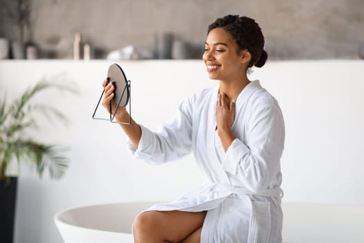 Joyful black woman in white bathrobe admiring her reflection in handheld mirror
