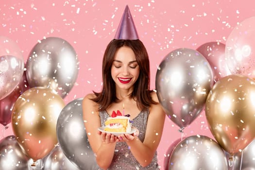 Delighted woman in a festive hat and sparkly dress admiring a slice of birthday cake