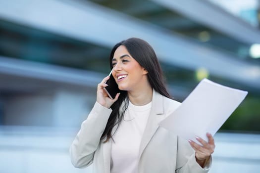 Smiling Latin entrepreneur lady chatting by phone standing outdoor